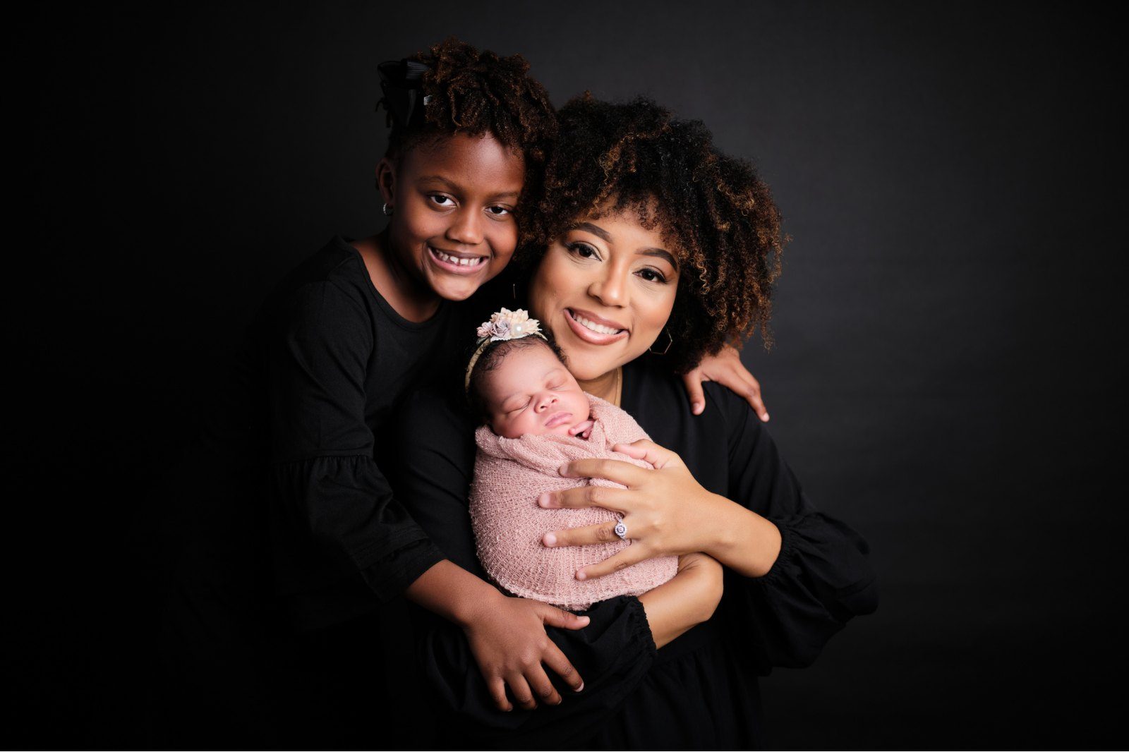 A mother holding a newborn with her daughter beside her against a black backdrop