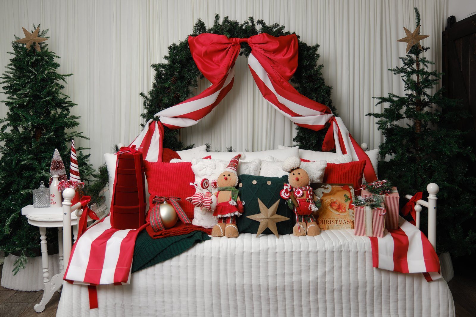 A white bed beneath a garland arch tied with a large red bow, flanked by two Christmas trees