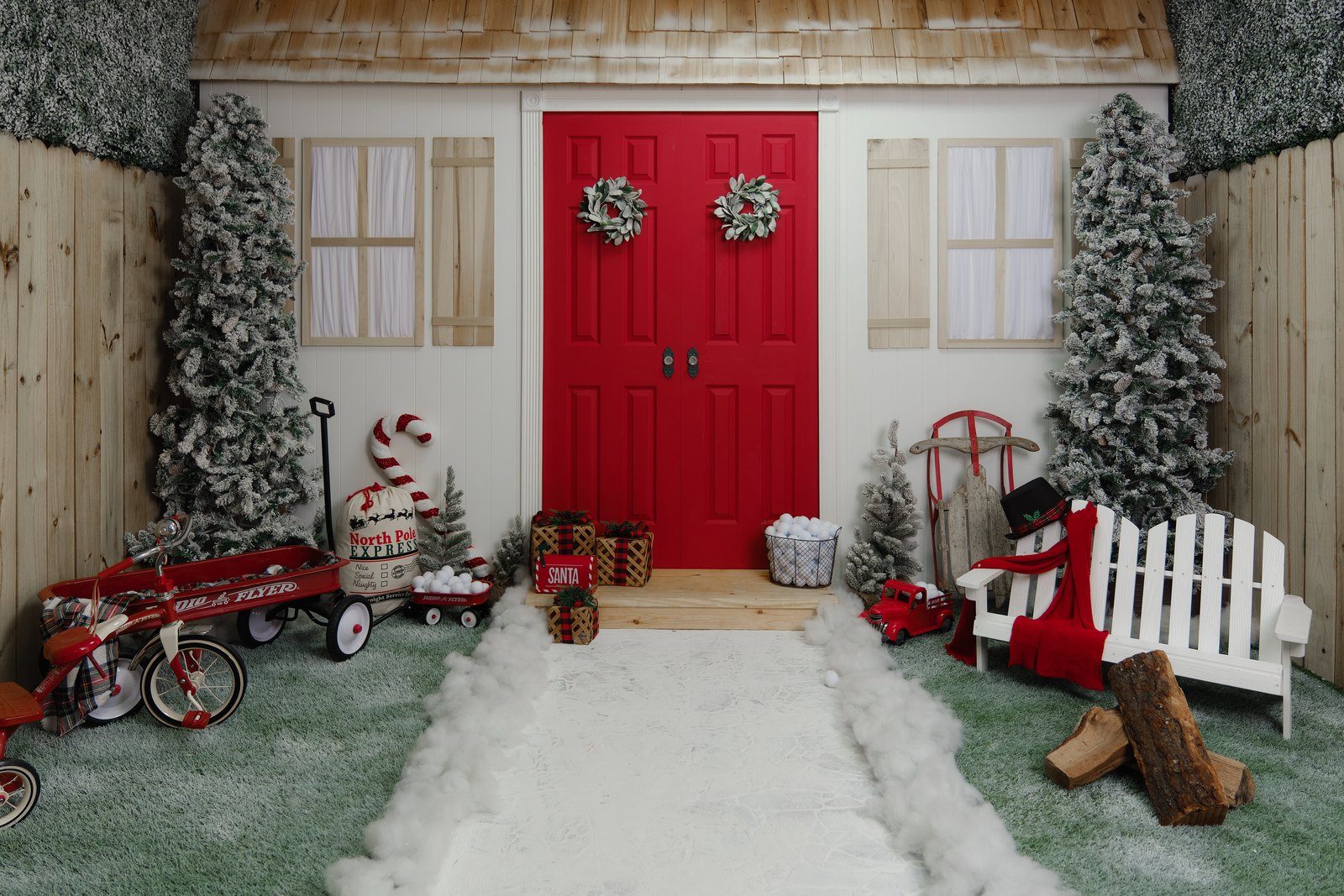 A cottage front with red double doors and wreaths, flocked trees, a snow path and a red wagon